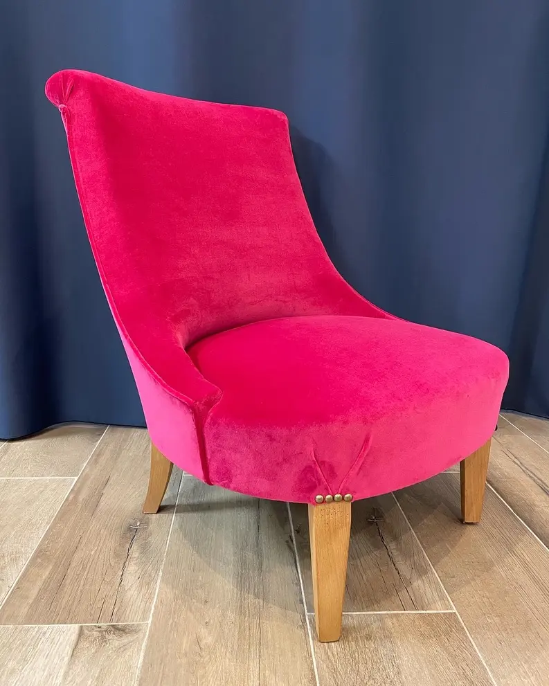 Pink upholstered accent chair with wooden legs on light wood floor against dark curtain.