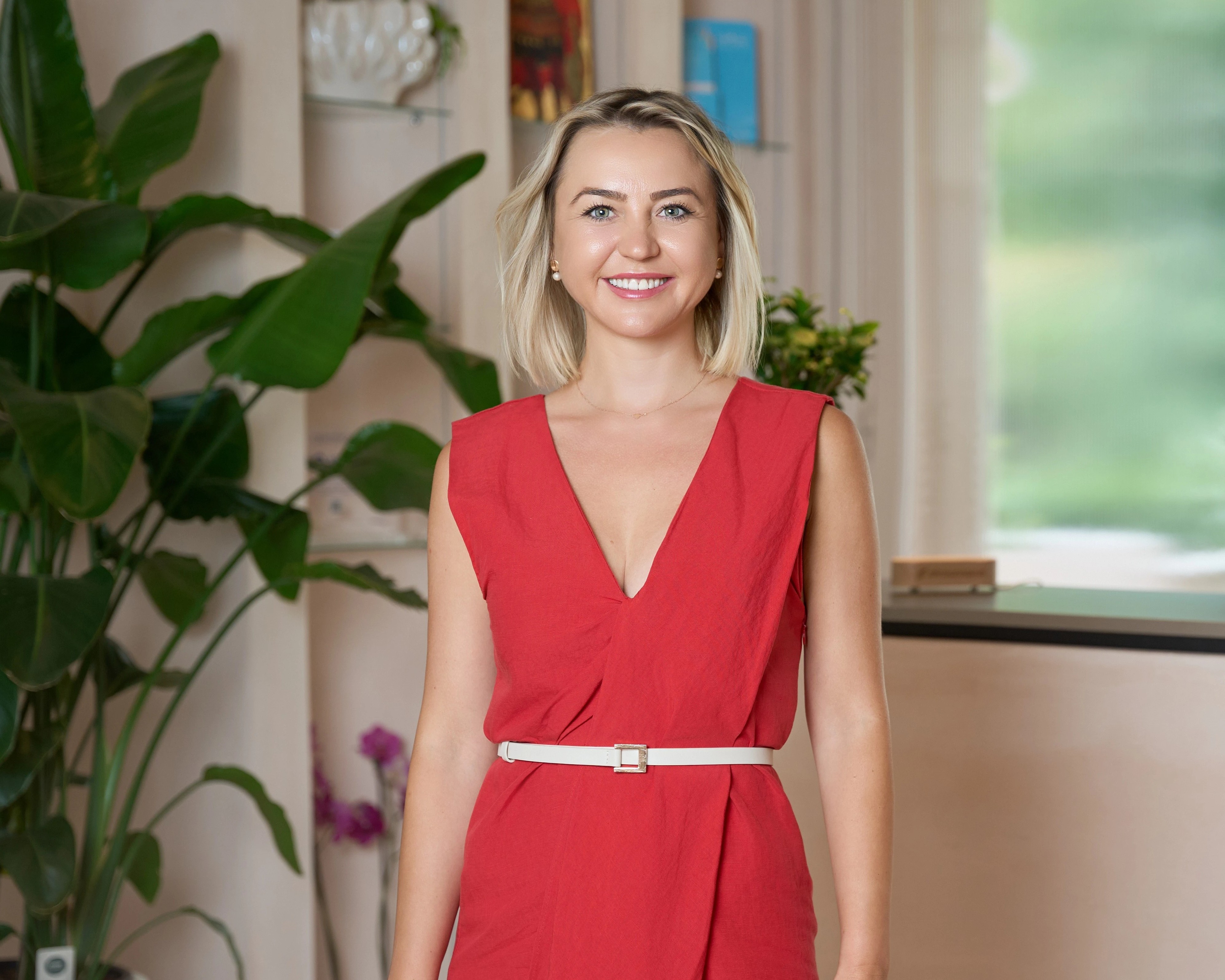 Smiling woman with blonde hair in a red sleeveless dress standing indoors near green plants.