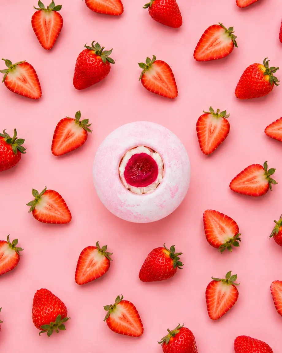 Pink strawberry bath bomb surrounded by fresh strawberries on a pink background