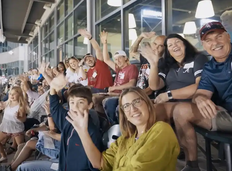 Group of spectators sitting in stadium seats waving and smiling at the camera.