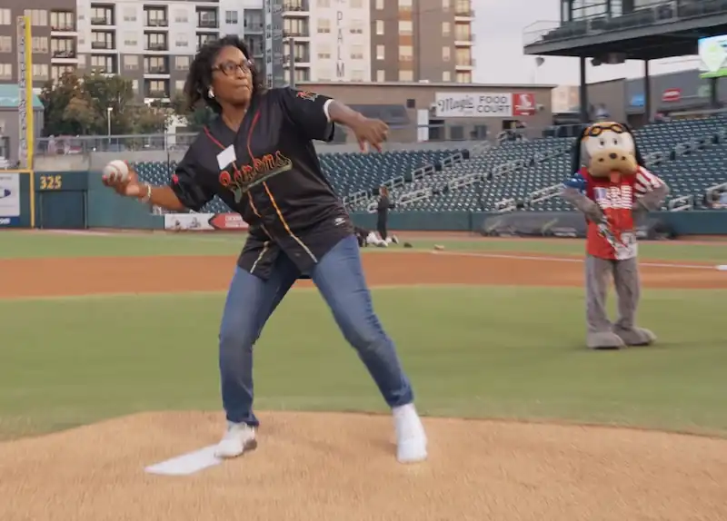 Woman wearing glasses and a baseball jersey throws the ceremonial first pitch on a baseball field with a mascot in background.