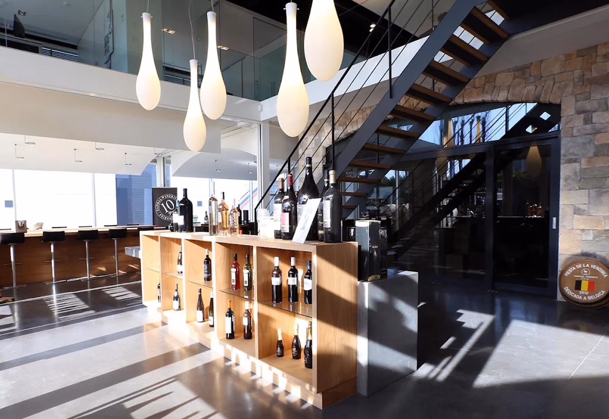 Moderne wijnwinkel met een houten displaykast gevuld met diverse wijnflessen, grote lichtgevende hanglampen erboven en een stalen trap met houten treden langs een stenen muur.
