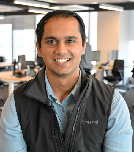 Smiling man with short dark hair wearing a light blue shirt and dark vest in an office setting.