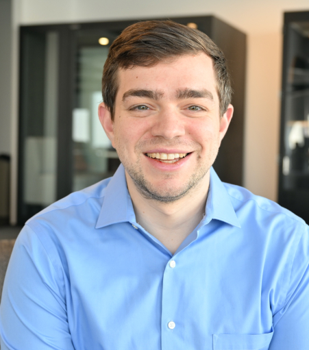 Smiling man with short brown hair wearing a light blue shirt indoors.