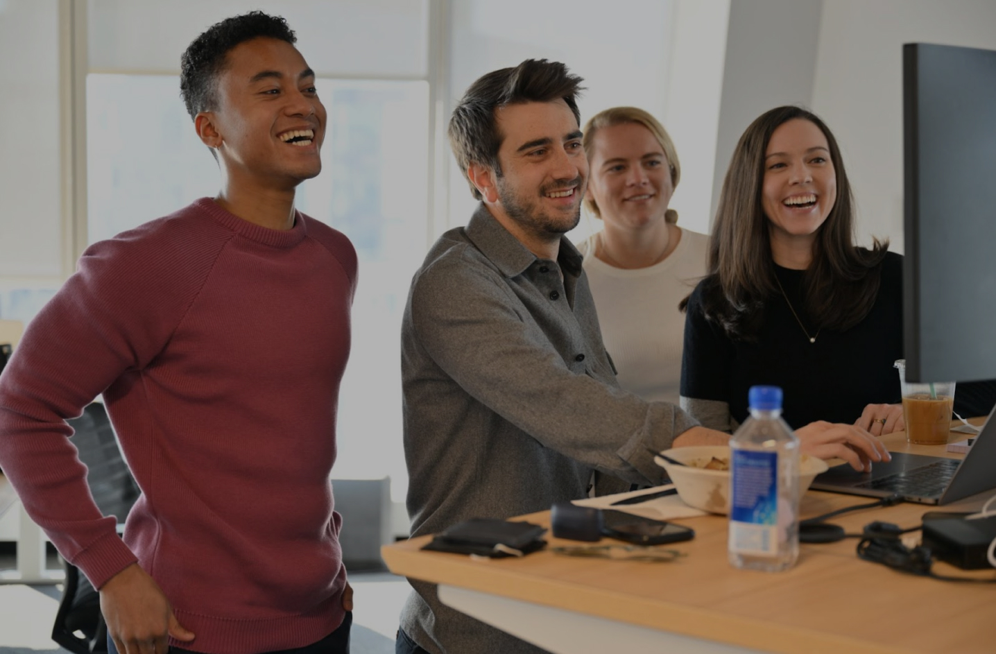 Four colleagues smiling and looking at a computer screen in an office with a standing desk.