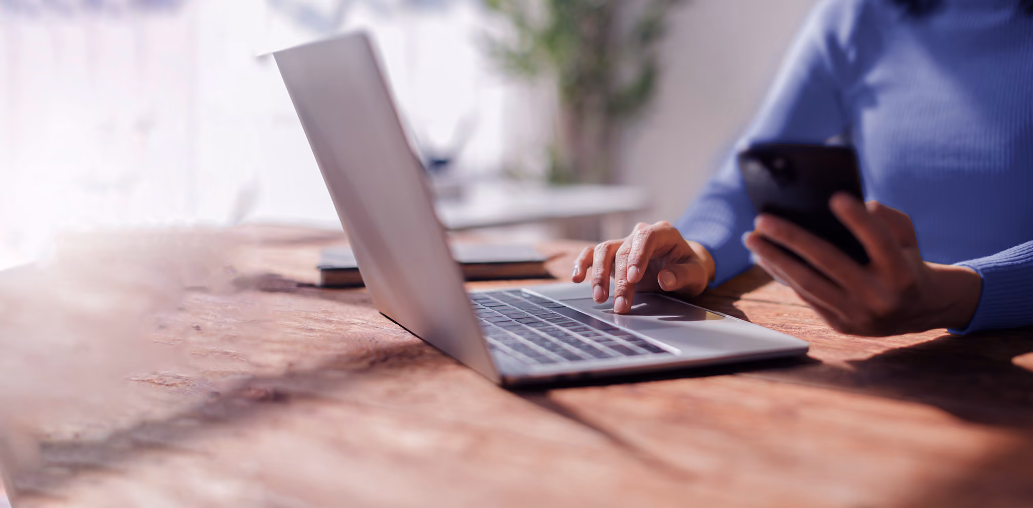 Person in blue sweater typing on laptop with one hand, holding phone in other hand on wooden table.