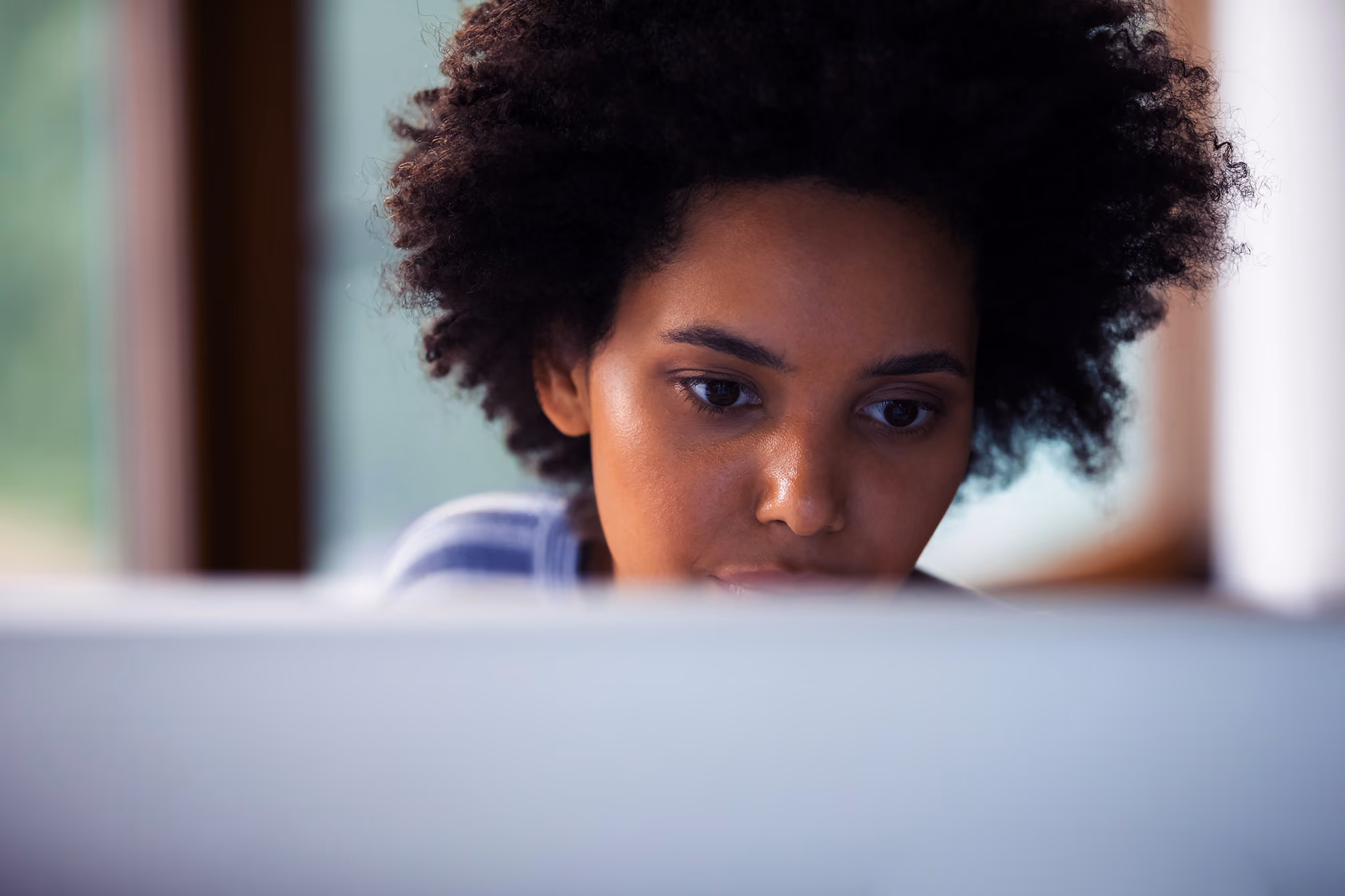 Woman with curly hair focused on a laptop screen with a blurred background.