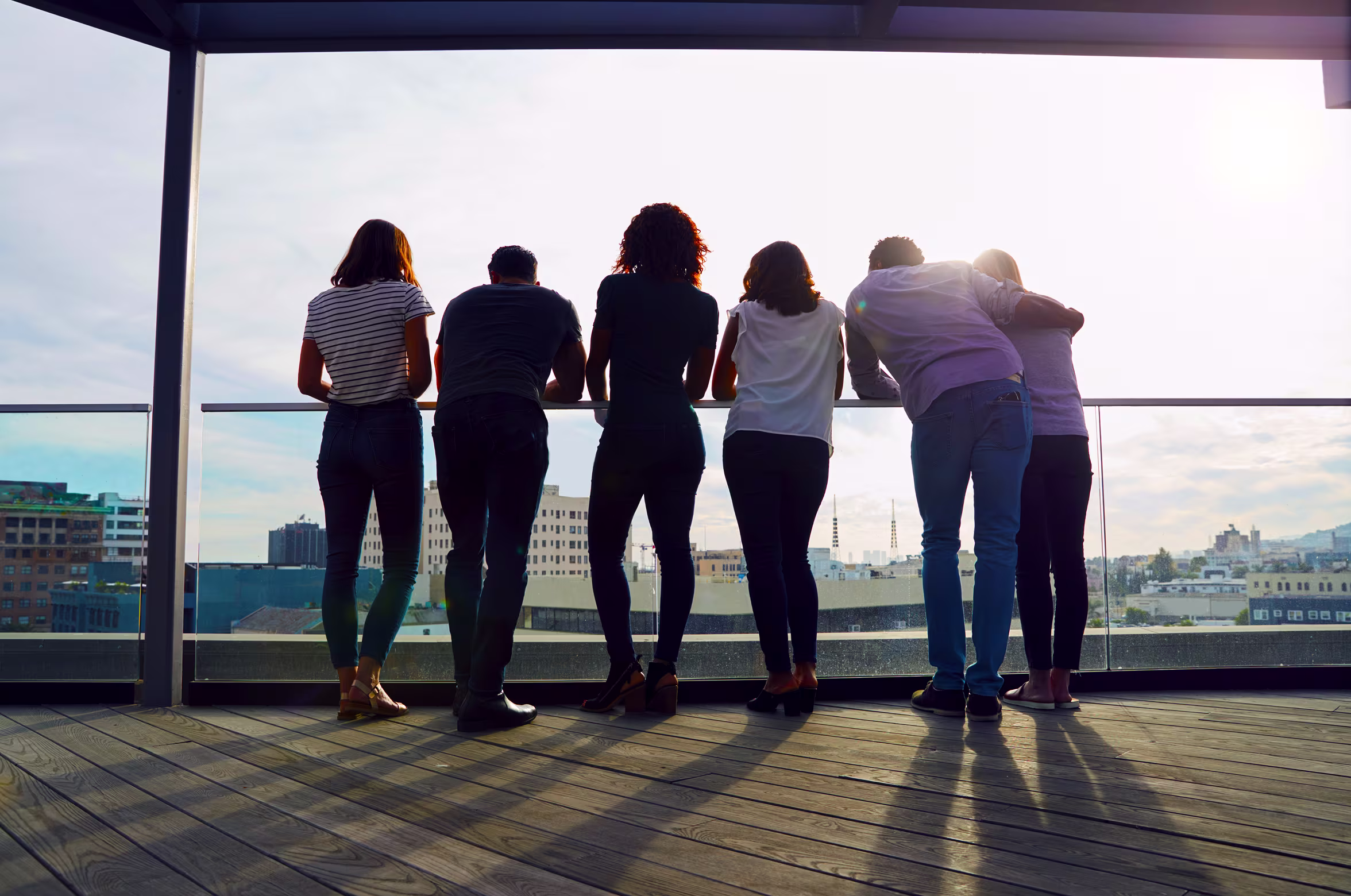 Group of six people standing and leaning on a rooftop railing, looking at the cityscape at sunset.