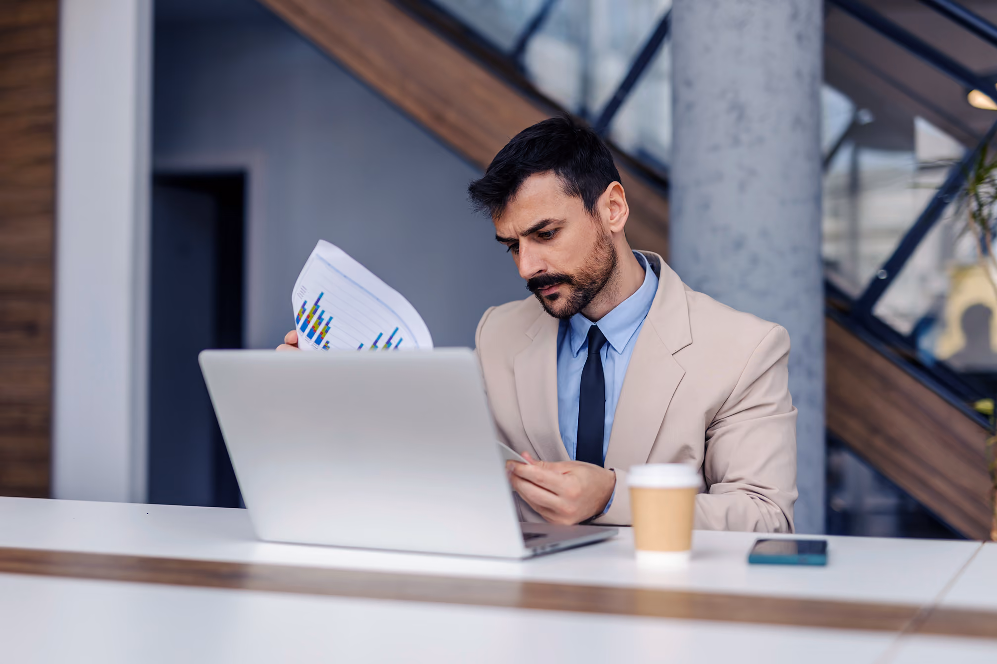 Man in beige suit reviewing a printed bar chart while working on a laptop at a desk with coffee and phone.