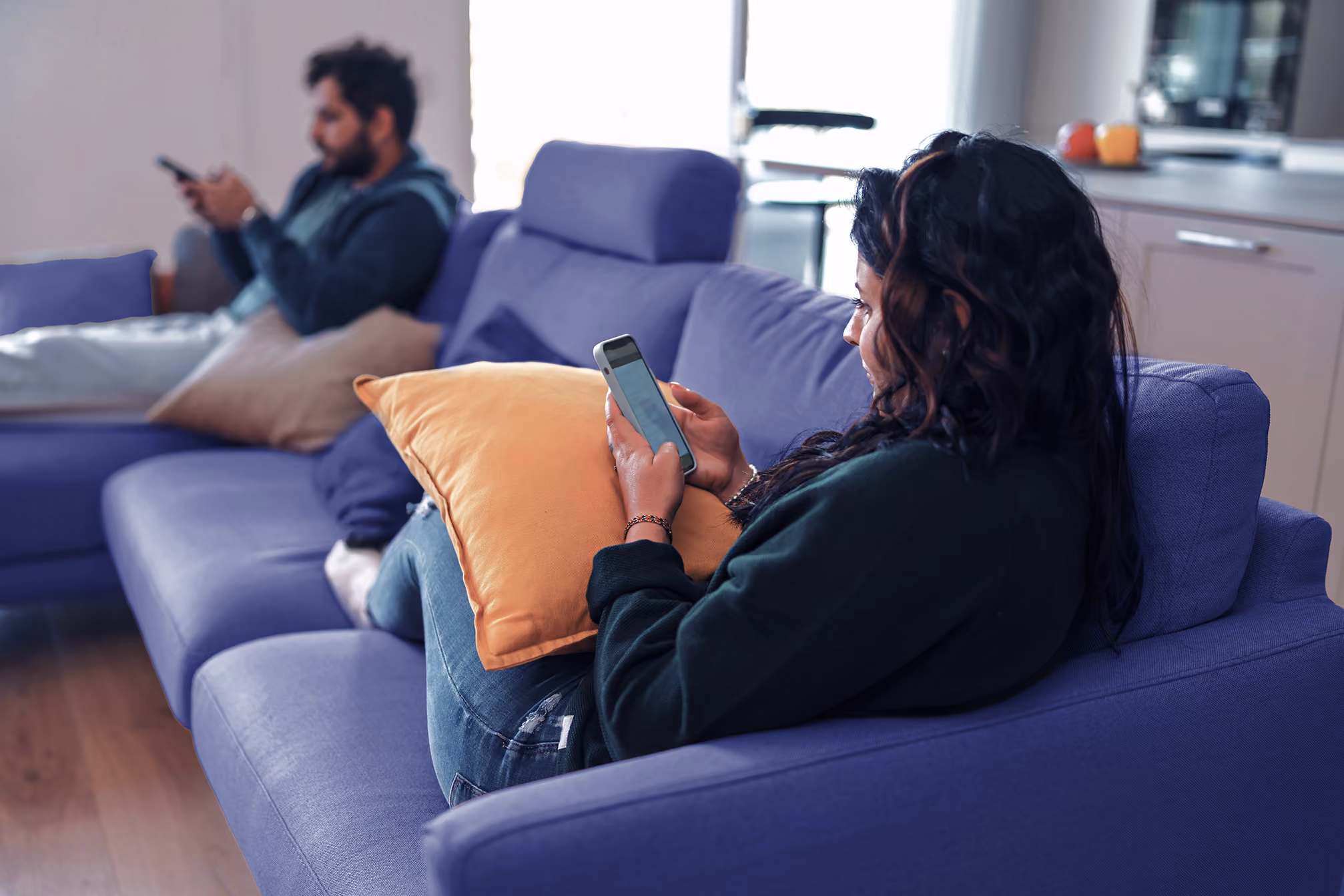 Two people sitting on a purple sofa using smartphones, woman holding an orange pillow.