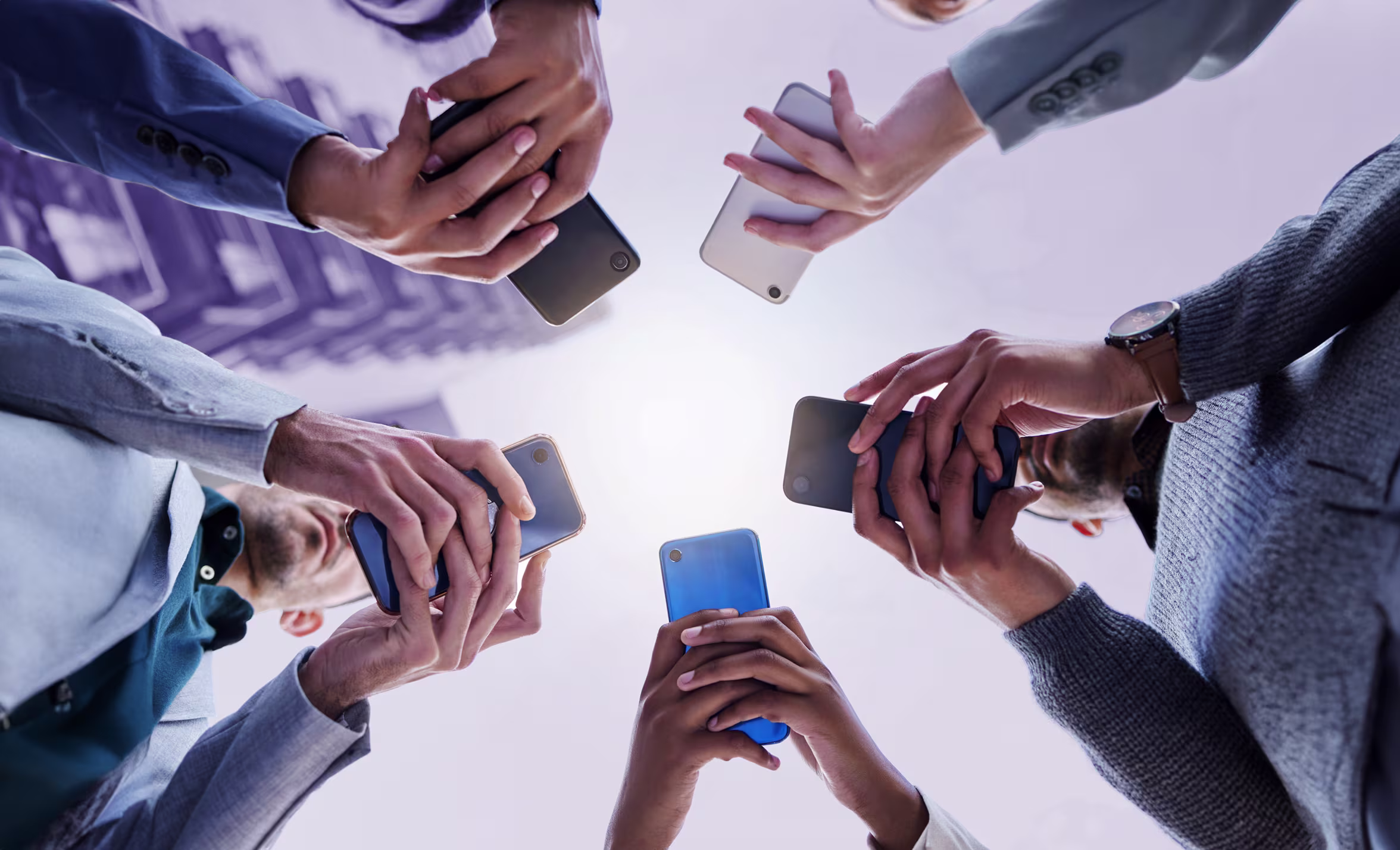 Group of diverse people standing in a circle looking down at their smartphones held in both hands.