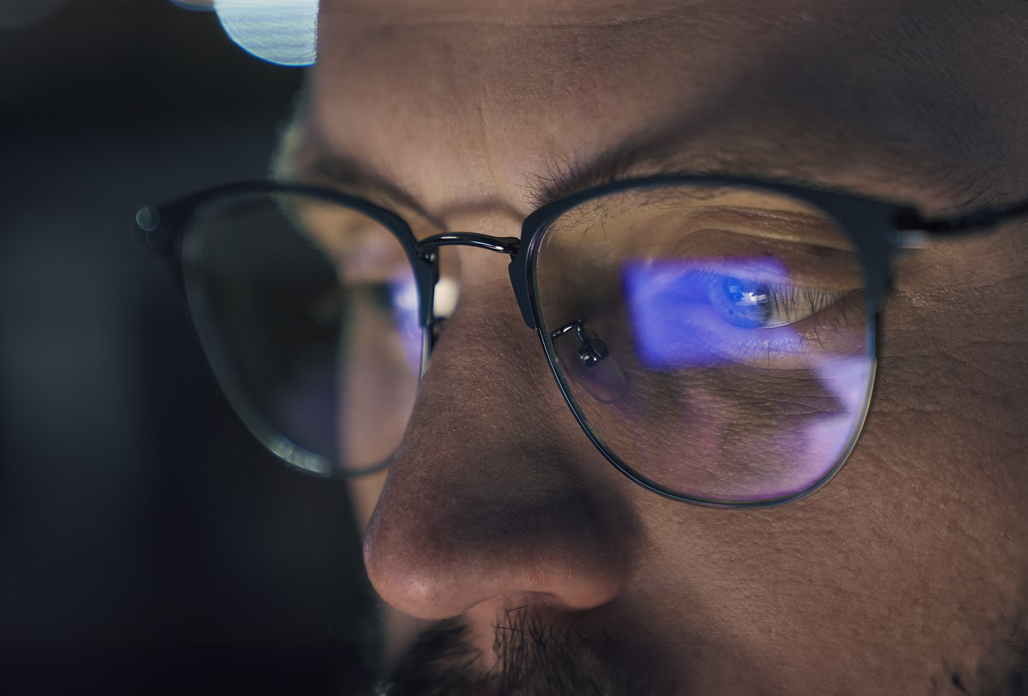 Close-up of a man wearing glasses reflecting a blue light from a screen.