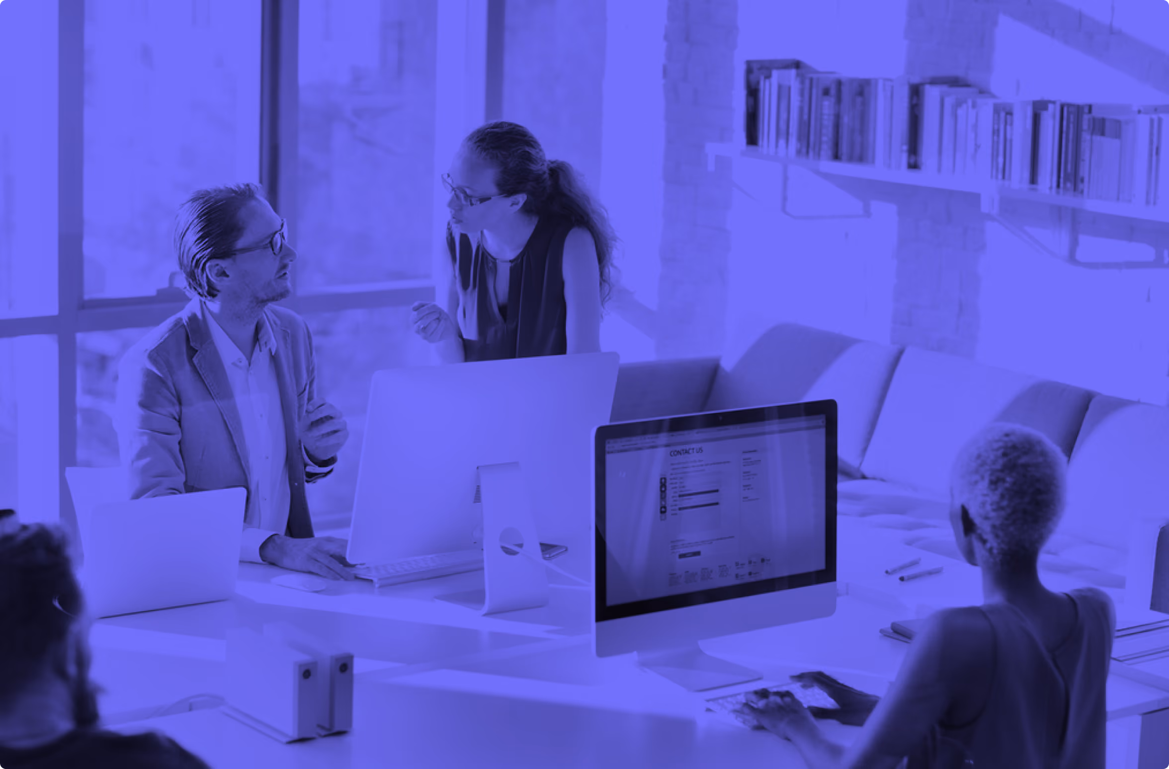 People working and discussing around computers in a modern office space with bookshelves.