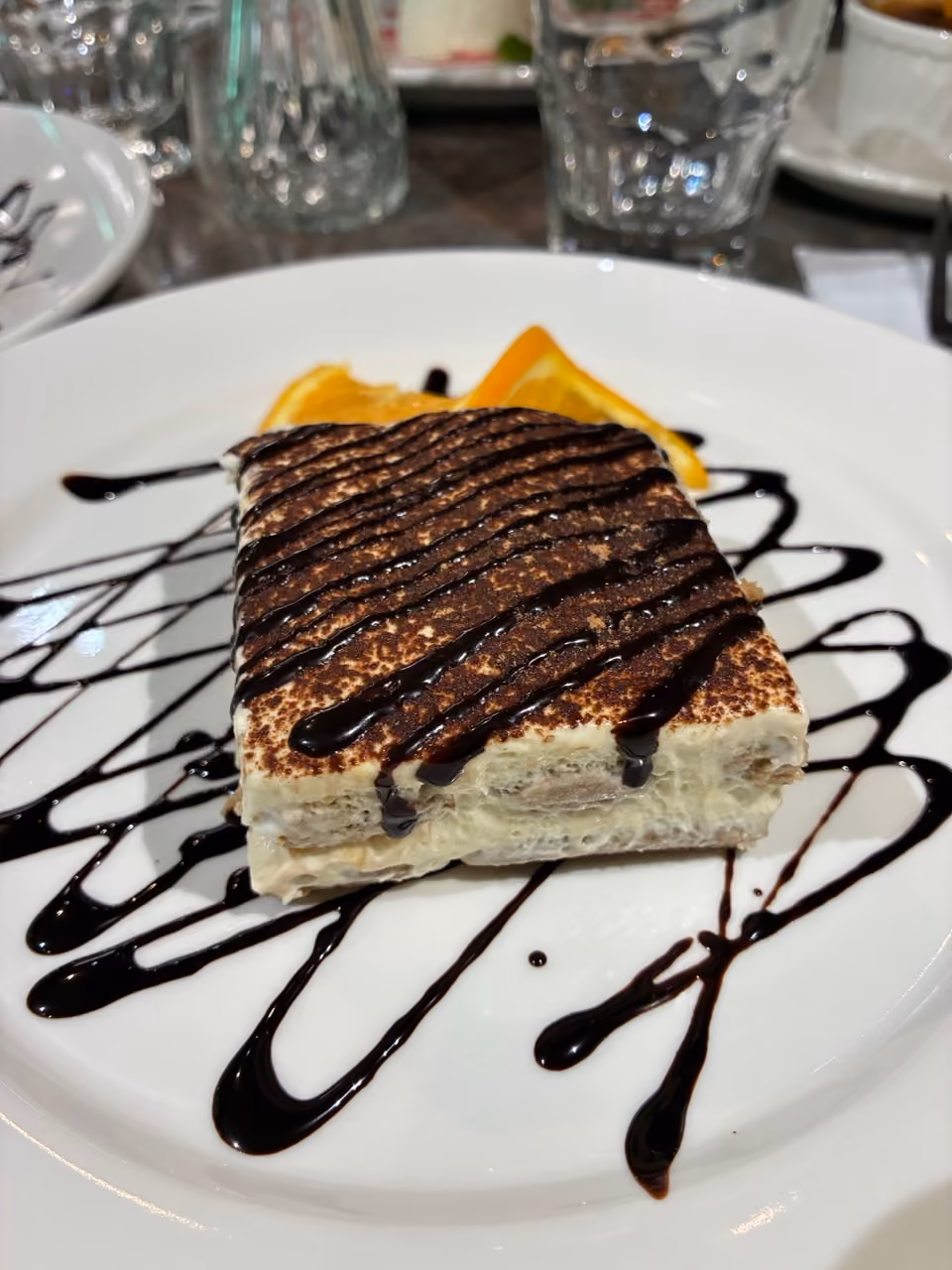 Square tiramisu cake drizzled with chocolate sauce on a white plate garnished with orange slices.
