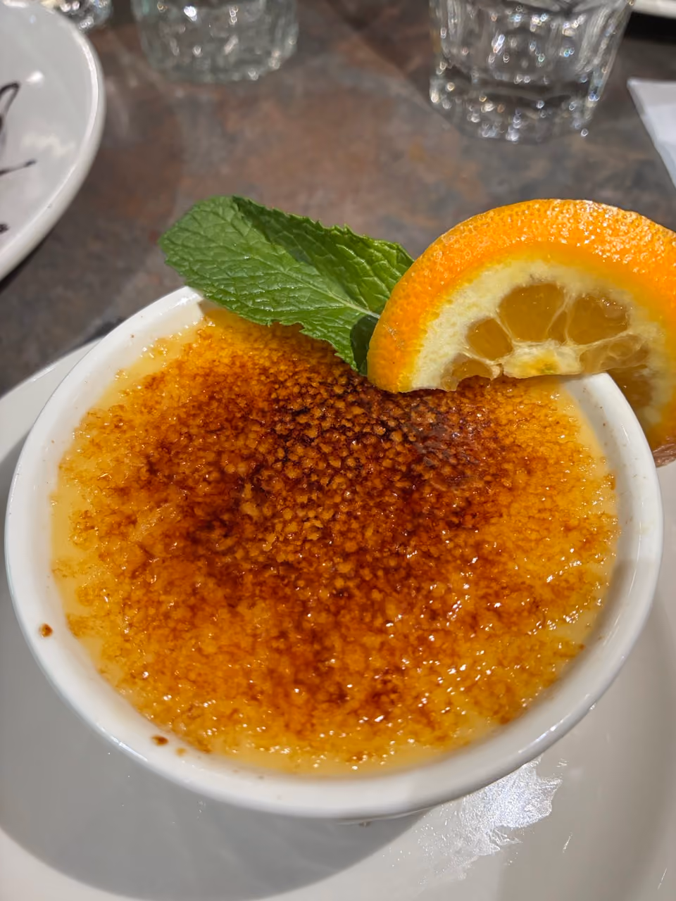 A crème brûlée dessert with caramelized sugar, garnished with a mint leaf and an orange slice.