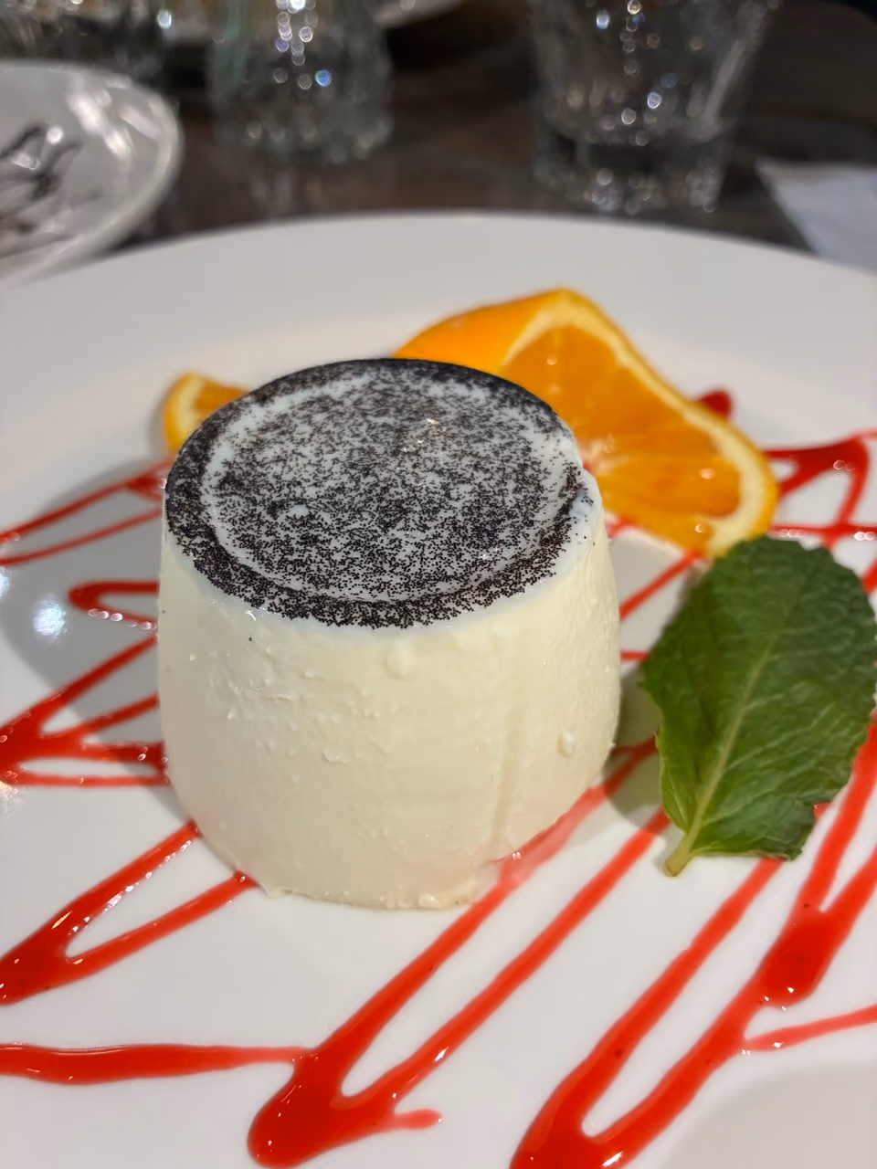 Creamy white dessert topped with black specks, served with orange slices, mint leaf, and red sauce drizzle.