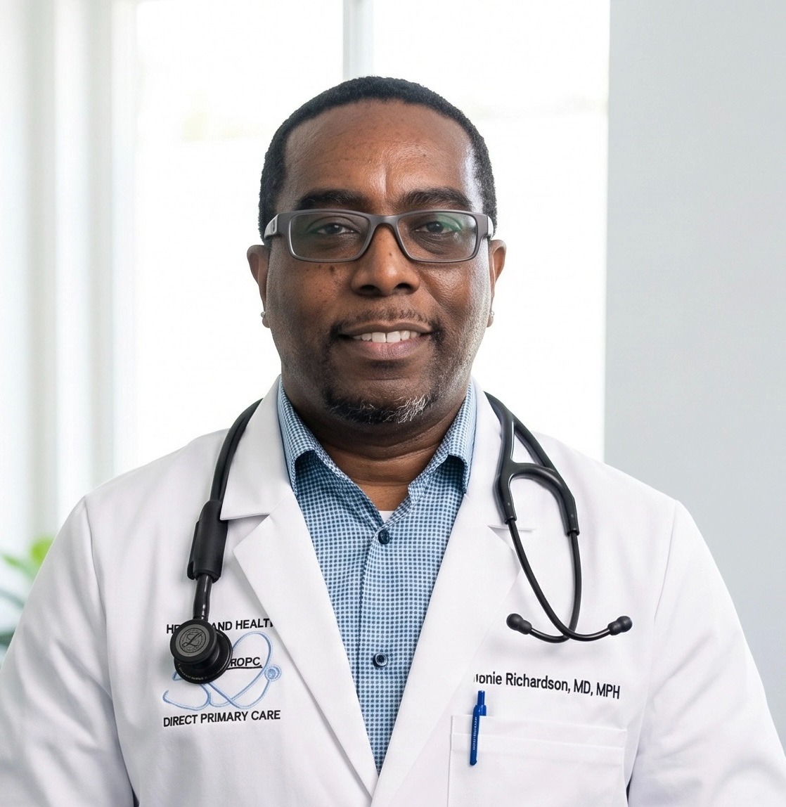 Smiling male doctor with glasses and stethoscope wearing a white coat and blue checkered shirt indoors.