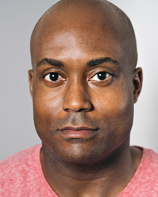 Close-up of a bald man with brown eyes wearing a red crew neck shirt looking straight ahead.