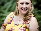 Smiling woman with braided blonde hair wearing a yellow floral dress outdoors.