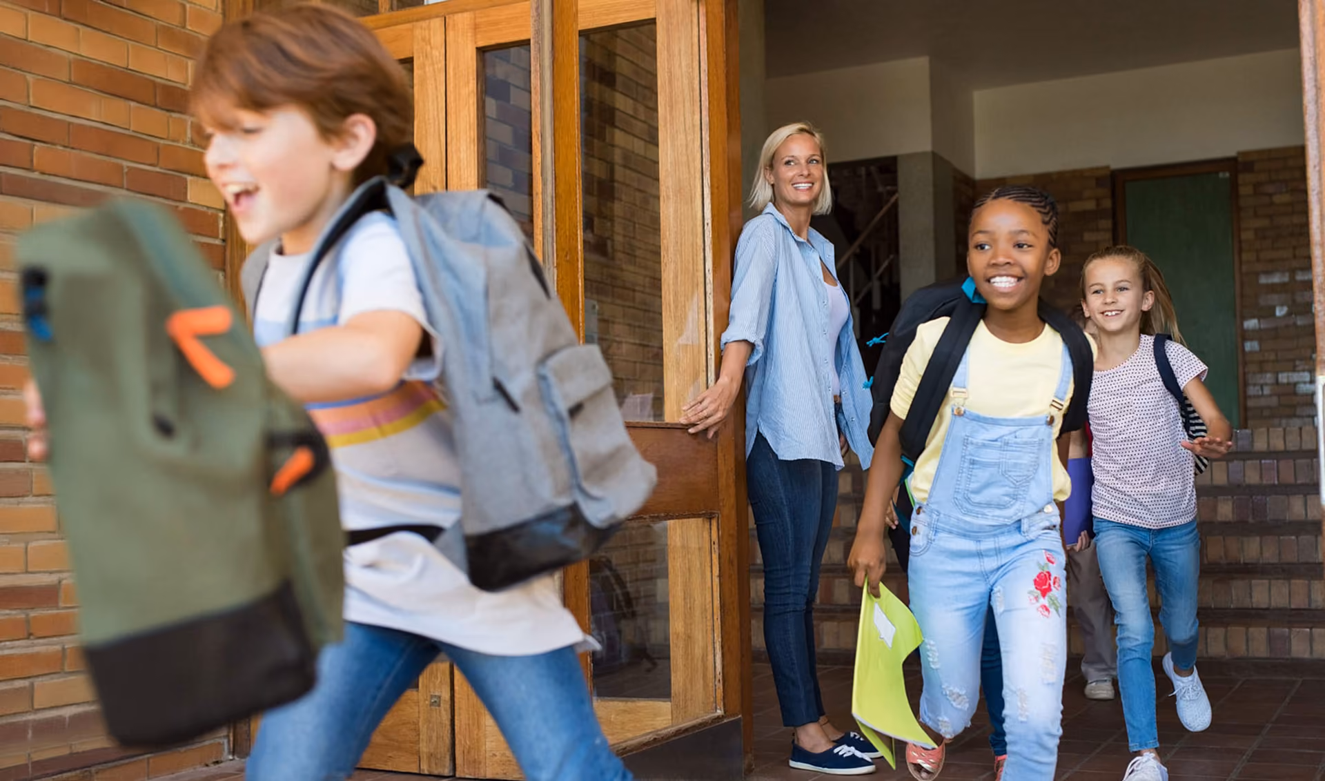Three children with backpacks happily leaving a school building, greeted by a smiling adult woman holding the door open.