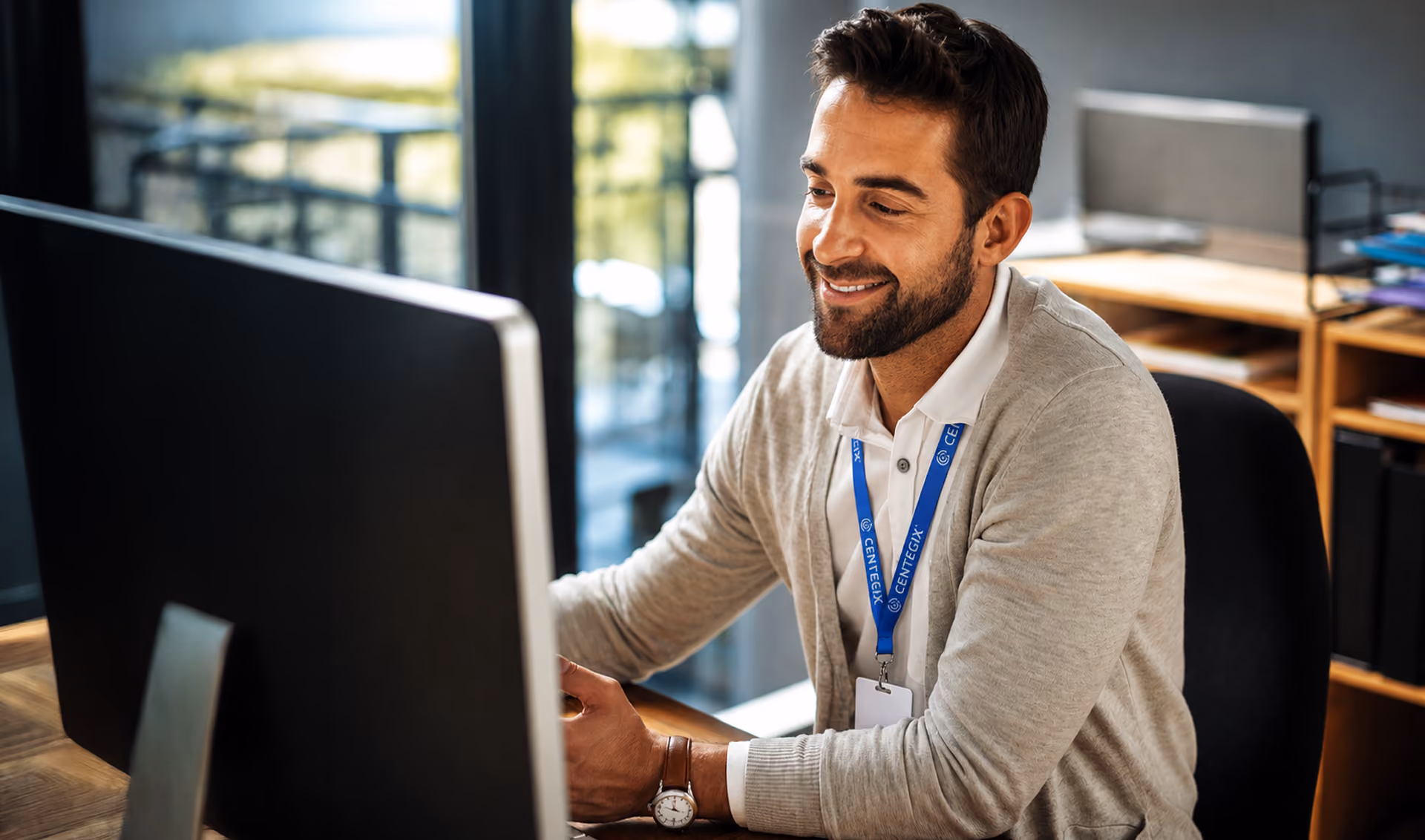 Smiling man with a beard sitting at a desk, looking at a computer monitor, wearing a beige cardigan and a blue lanyard in a bright office.