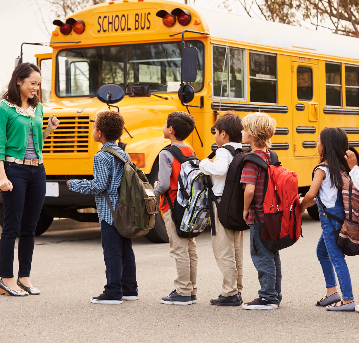 A woman smiling and greeting a line of six young children with backpacks standing on a pavement in front of a yellow school bus.