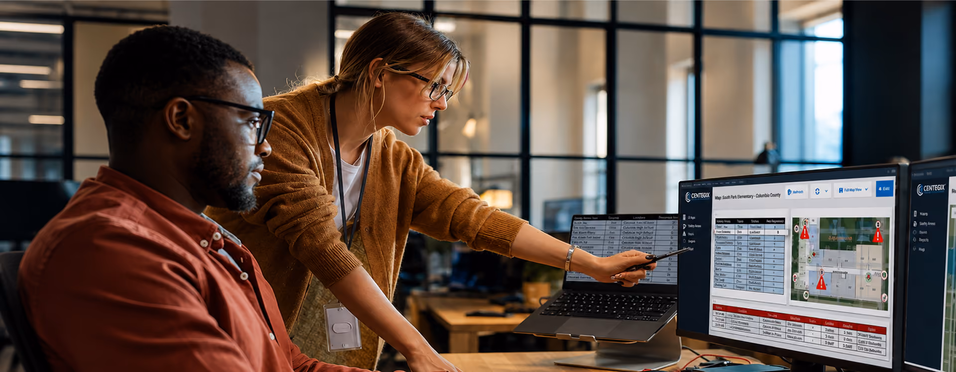 Two colleagues working together in an office; a woman is pointing at data on a large monitor showing maps and tables while a man watches attentively.
