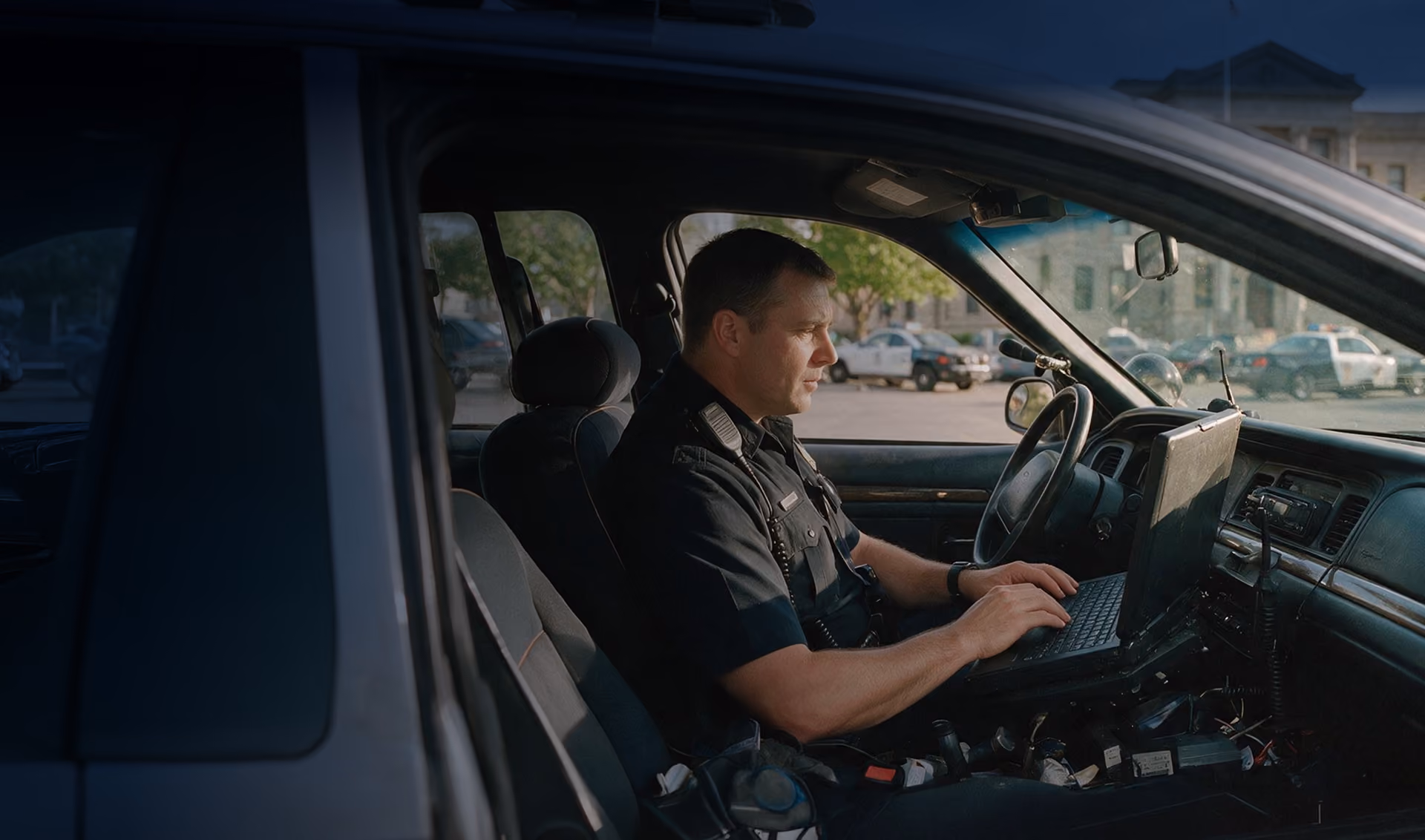 Police officer in uniform sitting in a patrol car using a laptop mounted on the center console with a parking lot visible outside the car window.