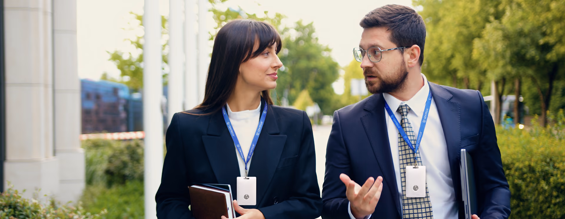 Two business professionals, a woman and a man, wearing suits and ID badges, walking outside engaged in conversation while carrying folders and a tablet.