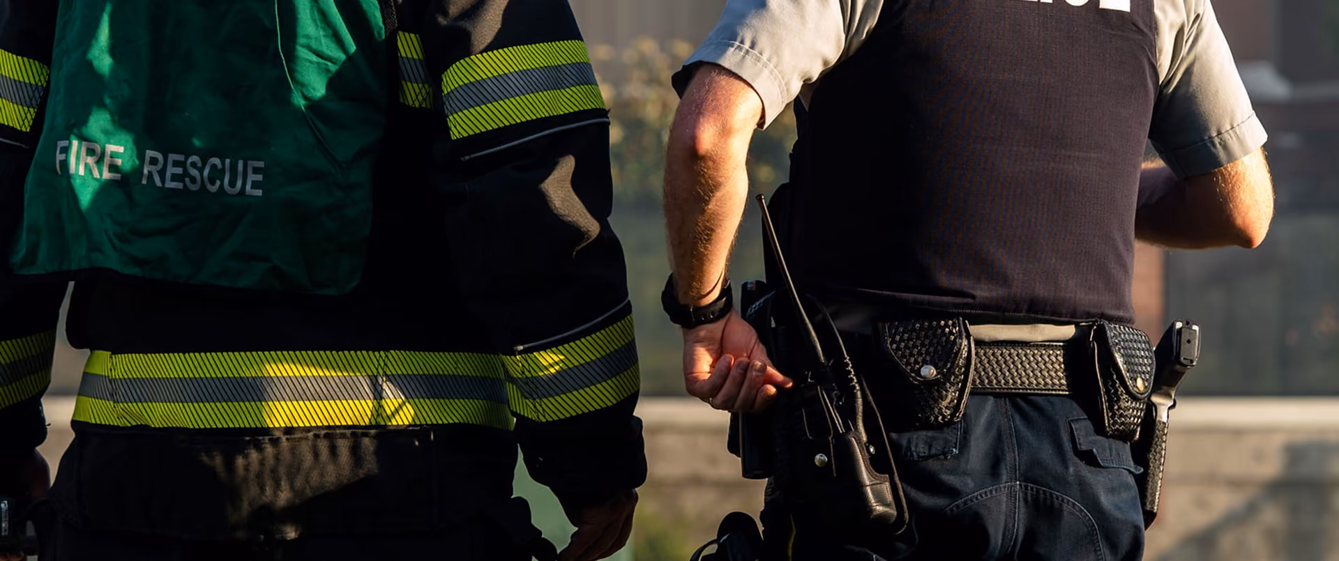 Back view of a firefighter wearing a green vest labeled 'FIRE RESCUE' standing next to a police officer in uniform with a radio, handcuffs, and a duty belt.