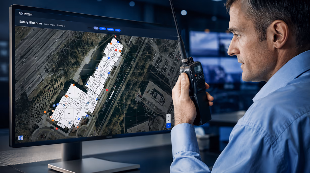 Man in a blue shirt holding a walkie-talkie and looking at a computer monitor displaying a detailed building safety blueprint overlayed on an aerial map.