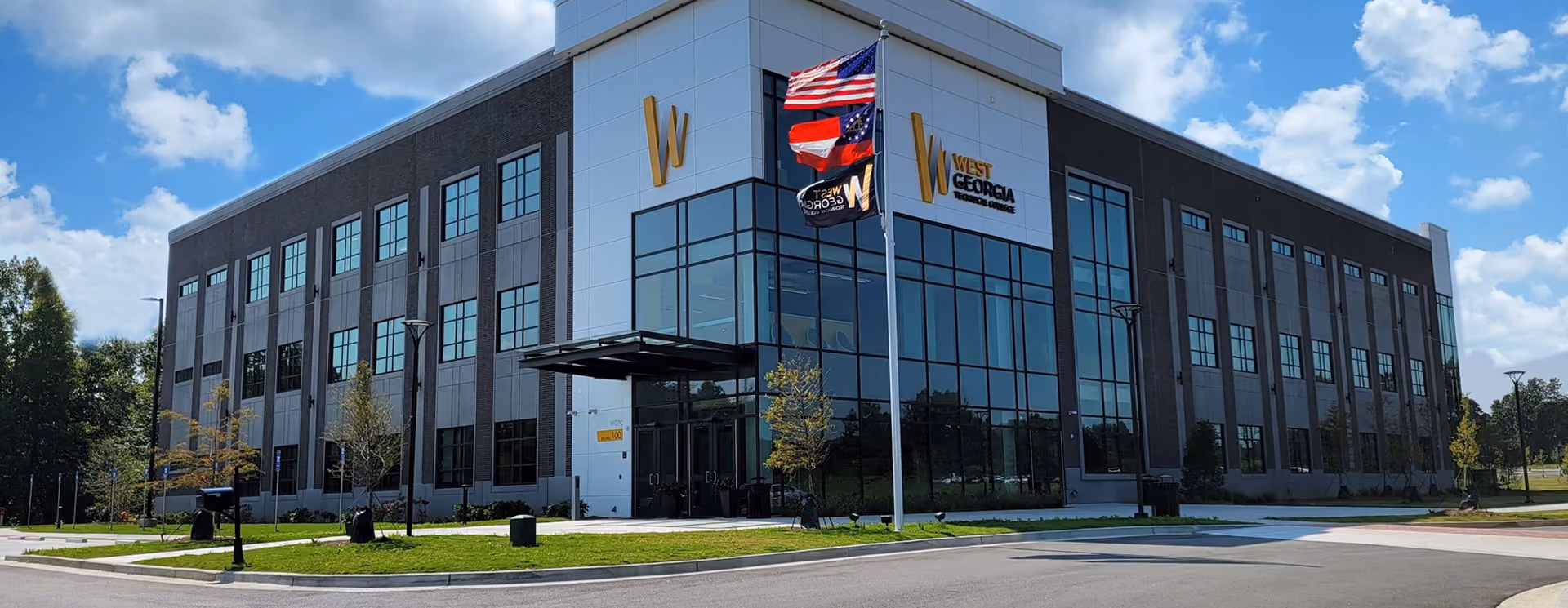 Modern three-story building with large glass windows and gray exterior panels, featuring signage for West Georgia, with American, Georgia state, and West Georgia flags flying on flagpoles in front under a partly cloudy blue sky.