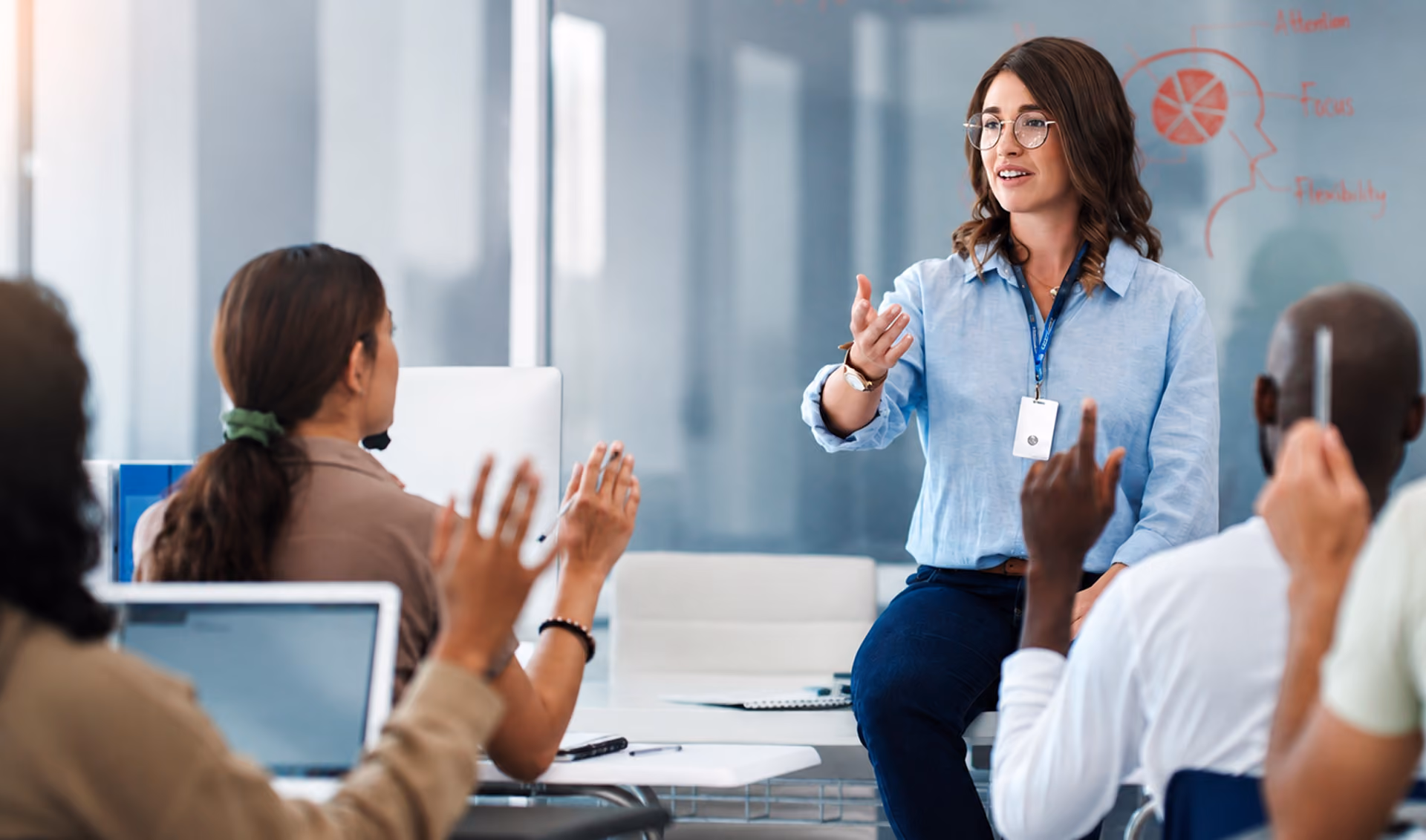 A woman wearing glasses and a blue shirt engages a group of diverse students in a modern classroom, with some students raising their hands to participate.