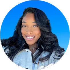 Smiling woman with long dark wavy hair wearing a light-colored shirt against a clear blue sky background.