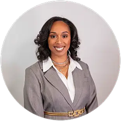 Portrait of a woman with medium-length dark curly hair wearing a light gray blazer over a white blouse and layered necklaces, smiling against a plain light gray background.