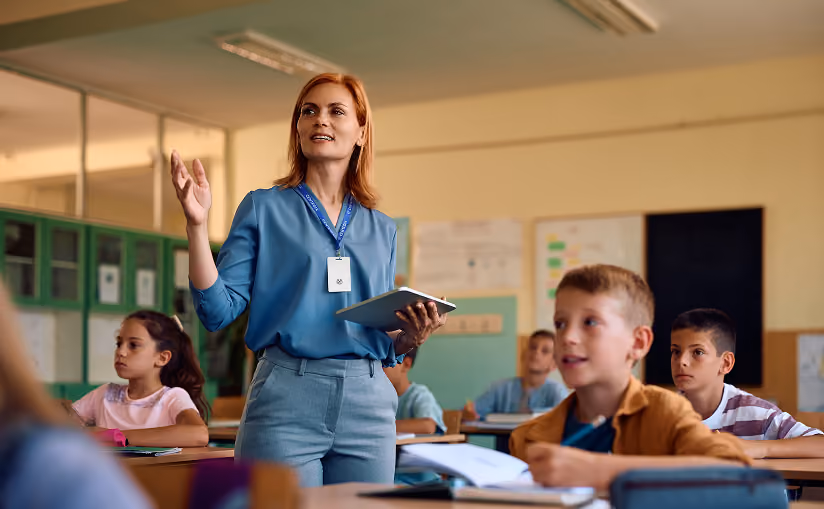 A female teacher holding a tablet and gesturing while standing in front of seated children in a classroom.