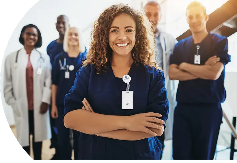 Smiling female healthcare professional in navy scrubs with arms crossed standing confidently in front of a diverse group of five medical staff in a bright hospital corridor.