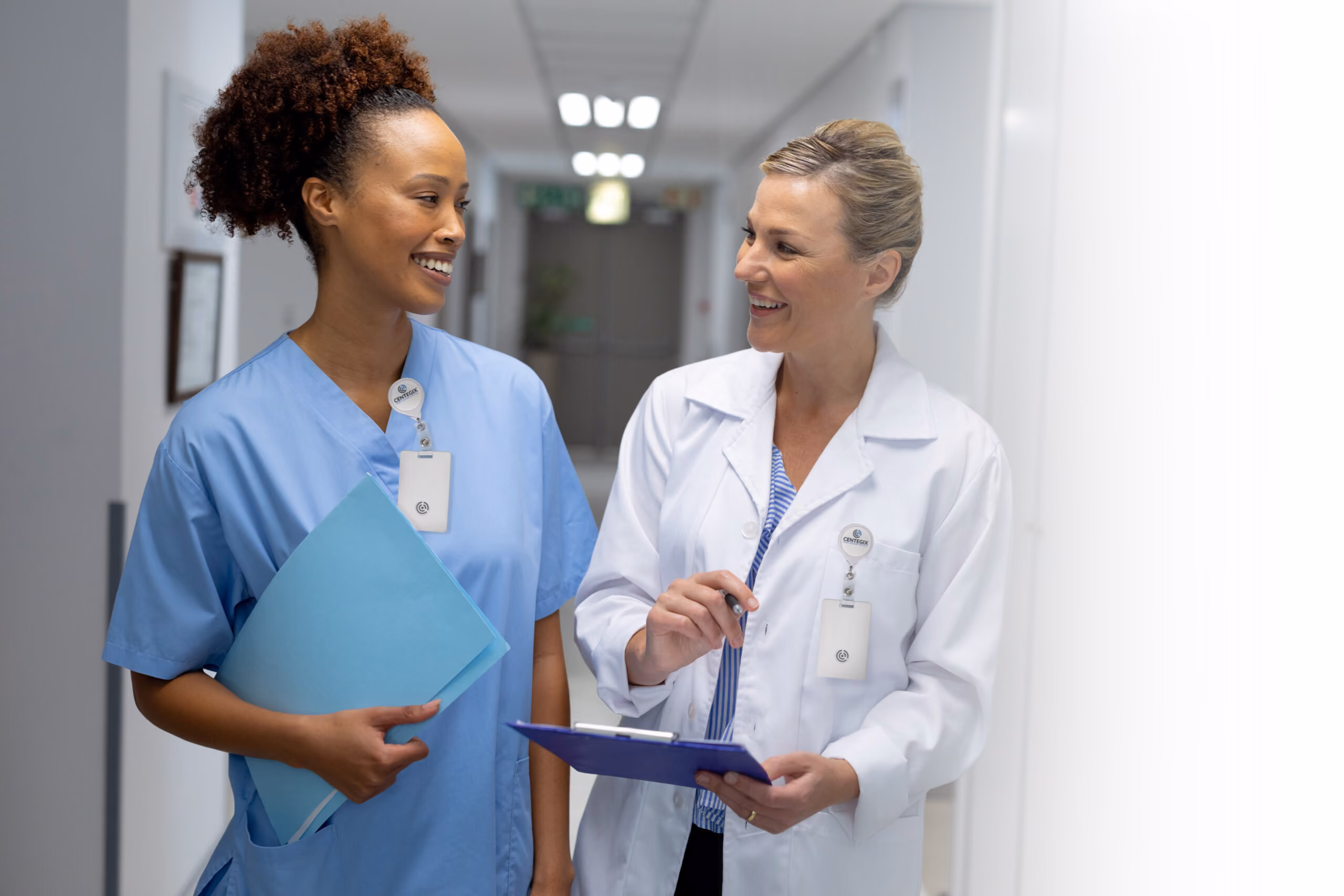 Two female healthcare professionals, one in blue scrubs holding a folder and the other in a white lab coat holding a clipboard, smiling and walking together in a hospital corridor.