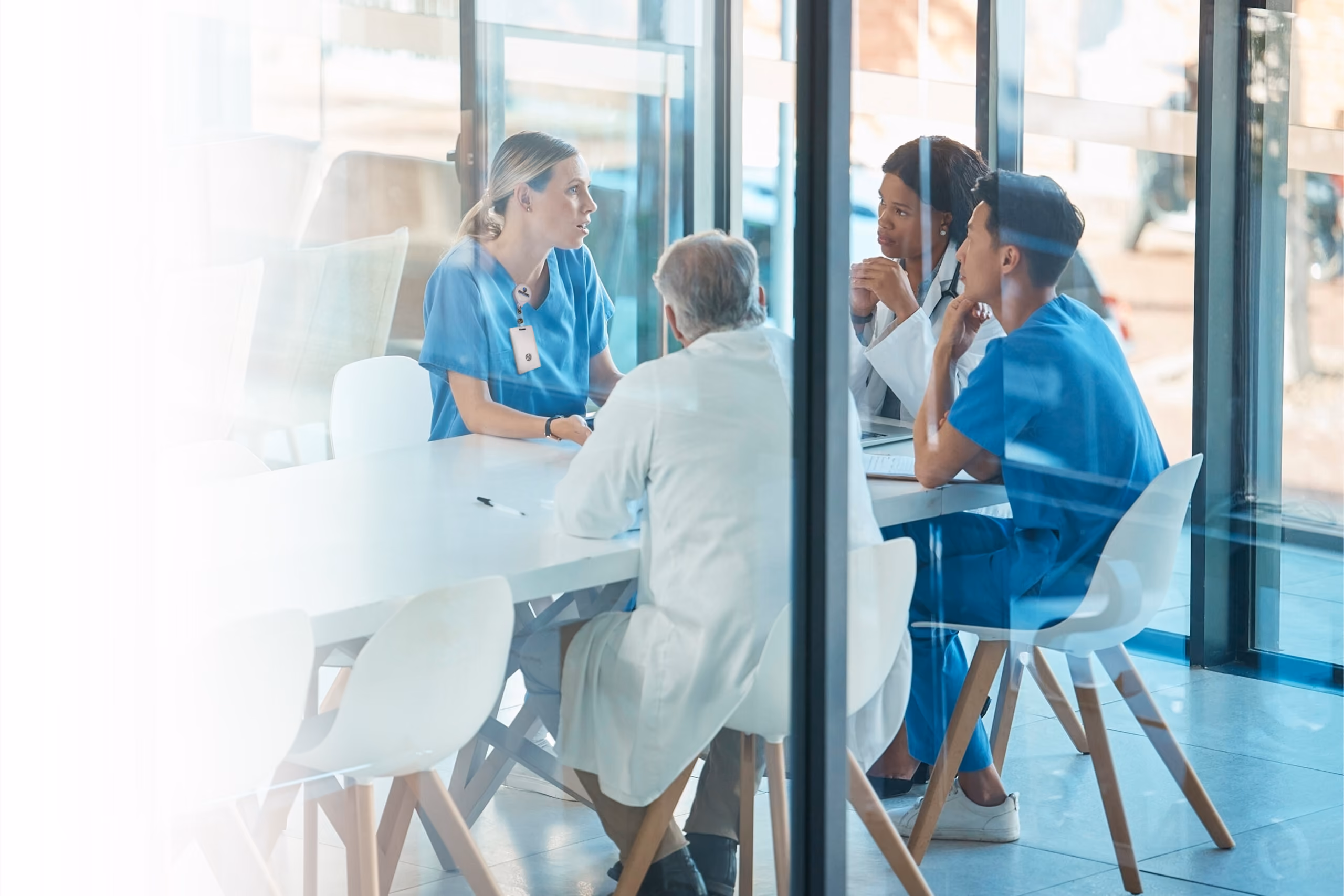 Four healthcare professionals in medical uniforms are sitting around a white table in a bright, modern meeting room having a discussion.