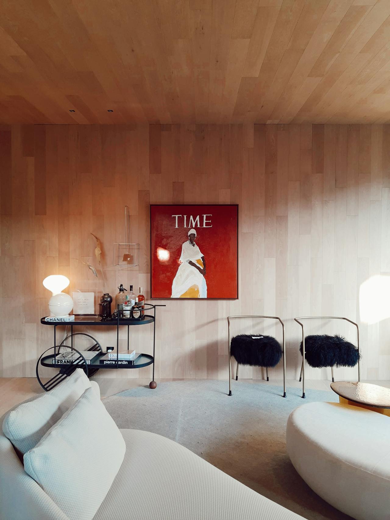 Modern living room with light wood panel walls, a white sofa, two black furry chairs, a round rug, and a bar cart under a framed red Time magazine cover.