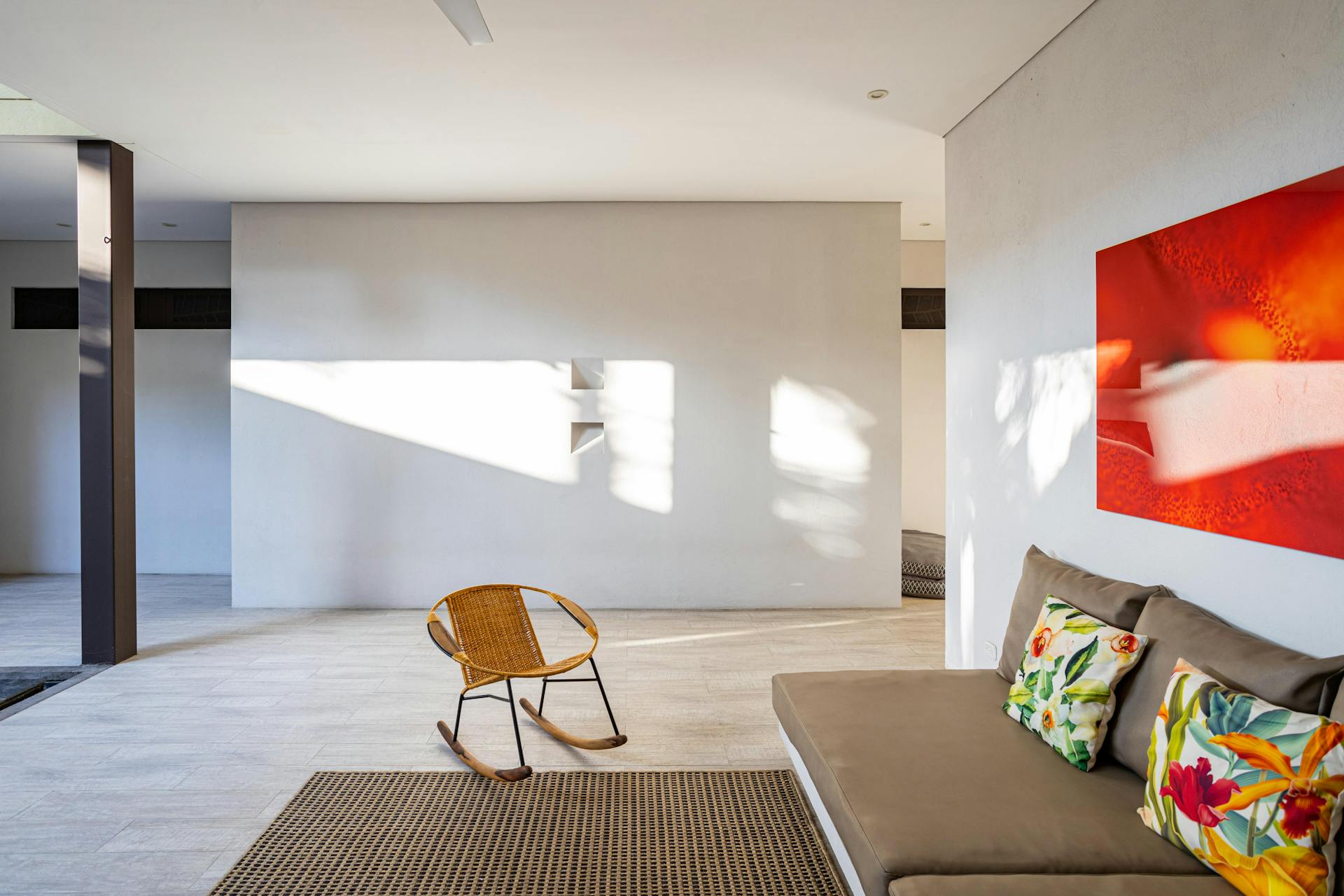 Bright modern room with a beige sofa adorned with floral pillows, a woven rocking chair, and a vibrant red abstract wall painting.