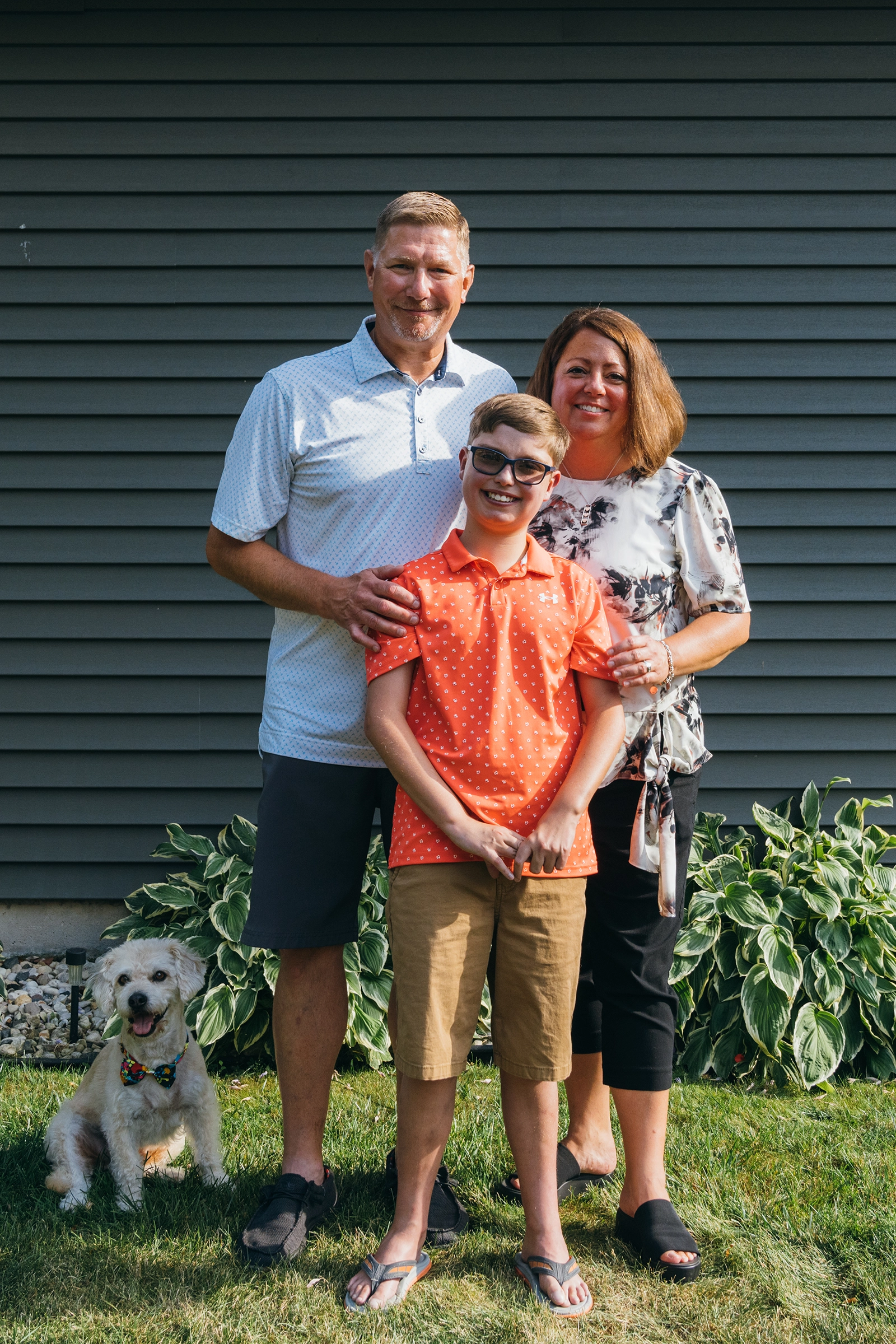 Smiling family of three standing outside near house siding with small dog wearing a colorful bow.