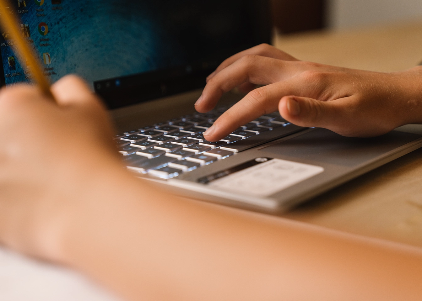 Close-up of a person's hand typing on a laptop keyboard with another hand holding a pencil.