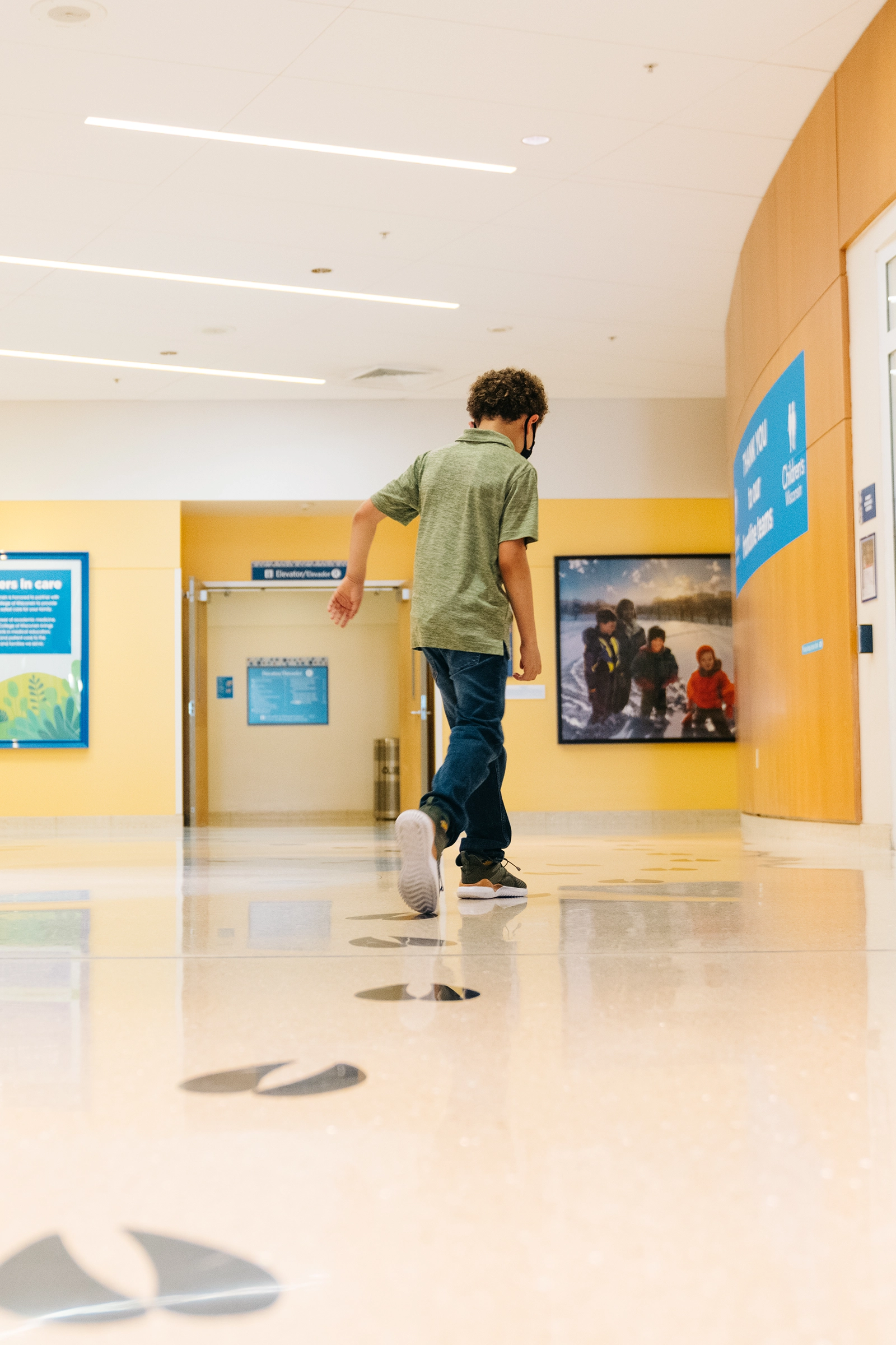 Child wearing a mask walking on floor decorated with large black footprints in a hospital corridor.