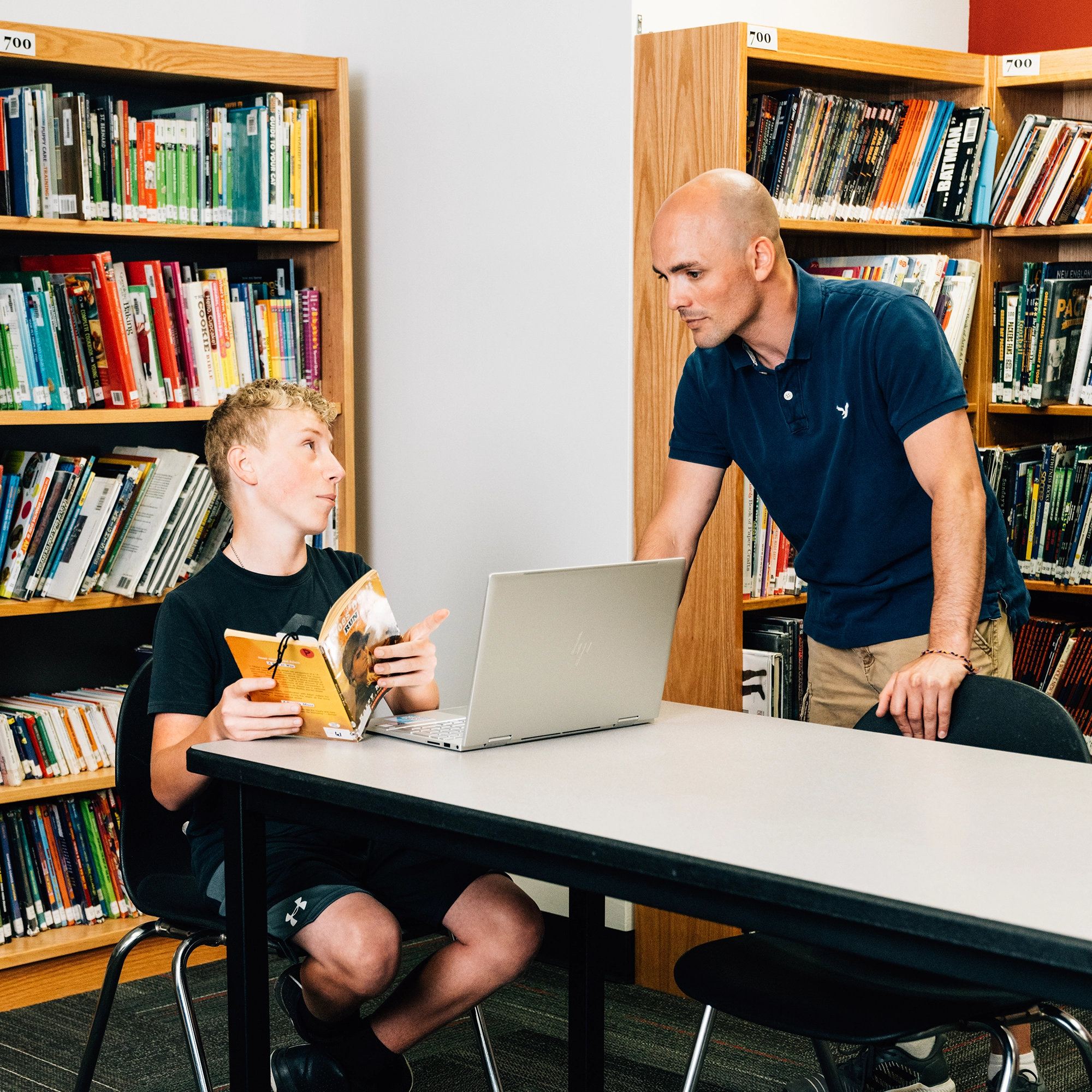 Teen boy sitting with a book and laptop talking to a man leaning on table in a library.