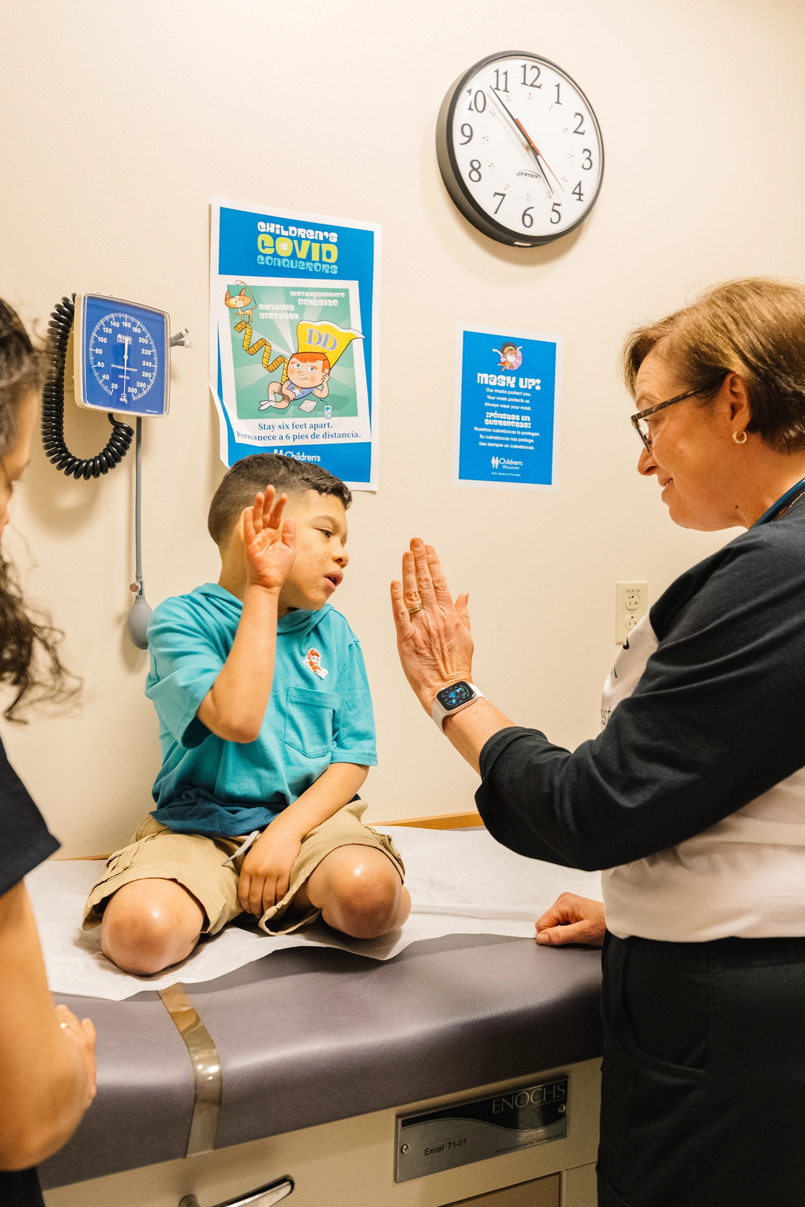 Child sitting on exam table giving a high-five to a smiling healthcare worker in an exam room.