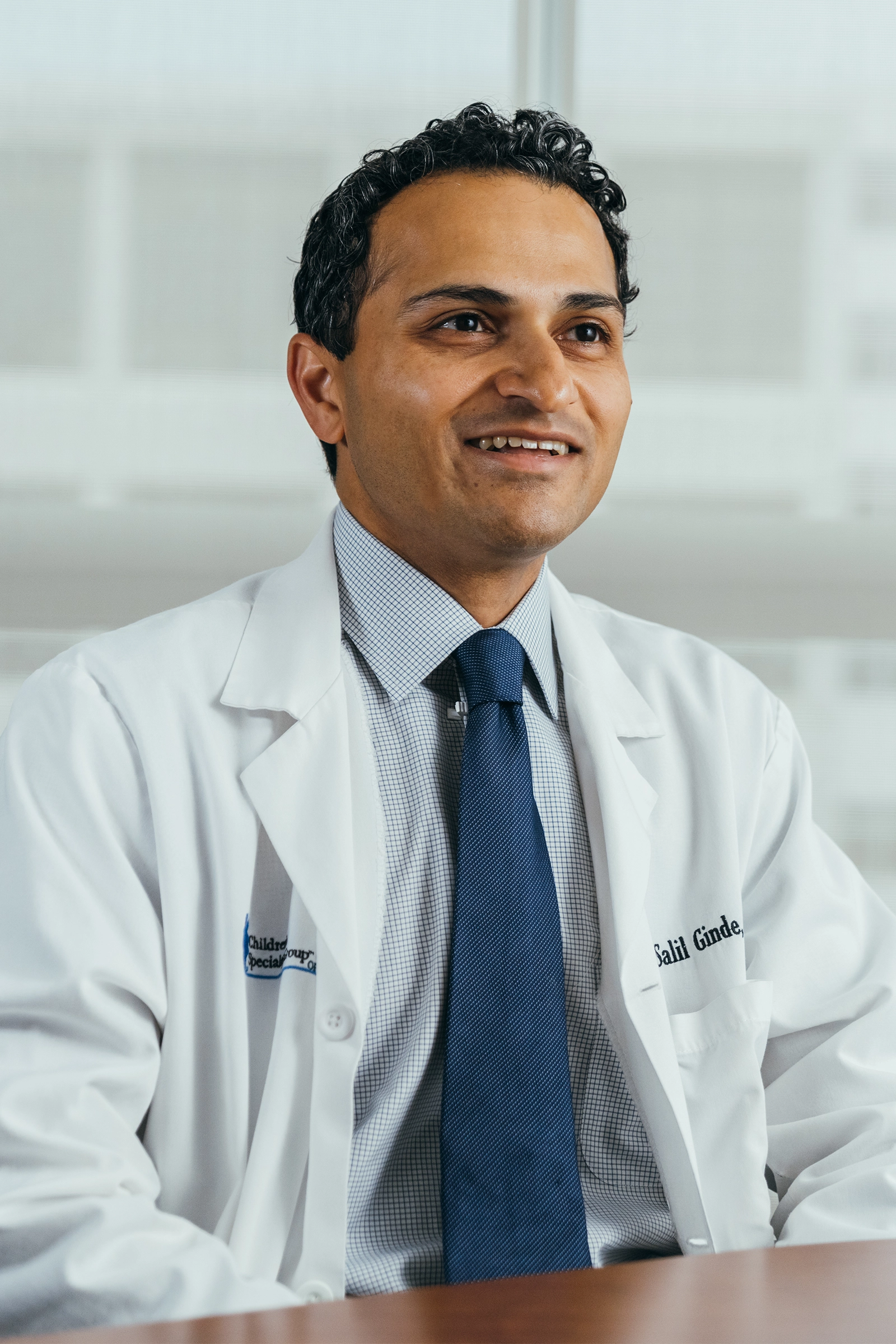 Smiling male doctor wearing a white coat, blue tie, and checkered shirt sitting at a desk.