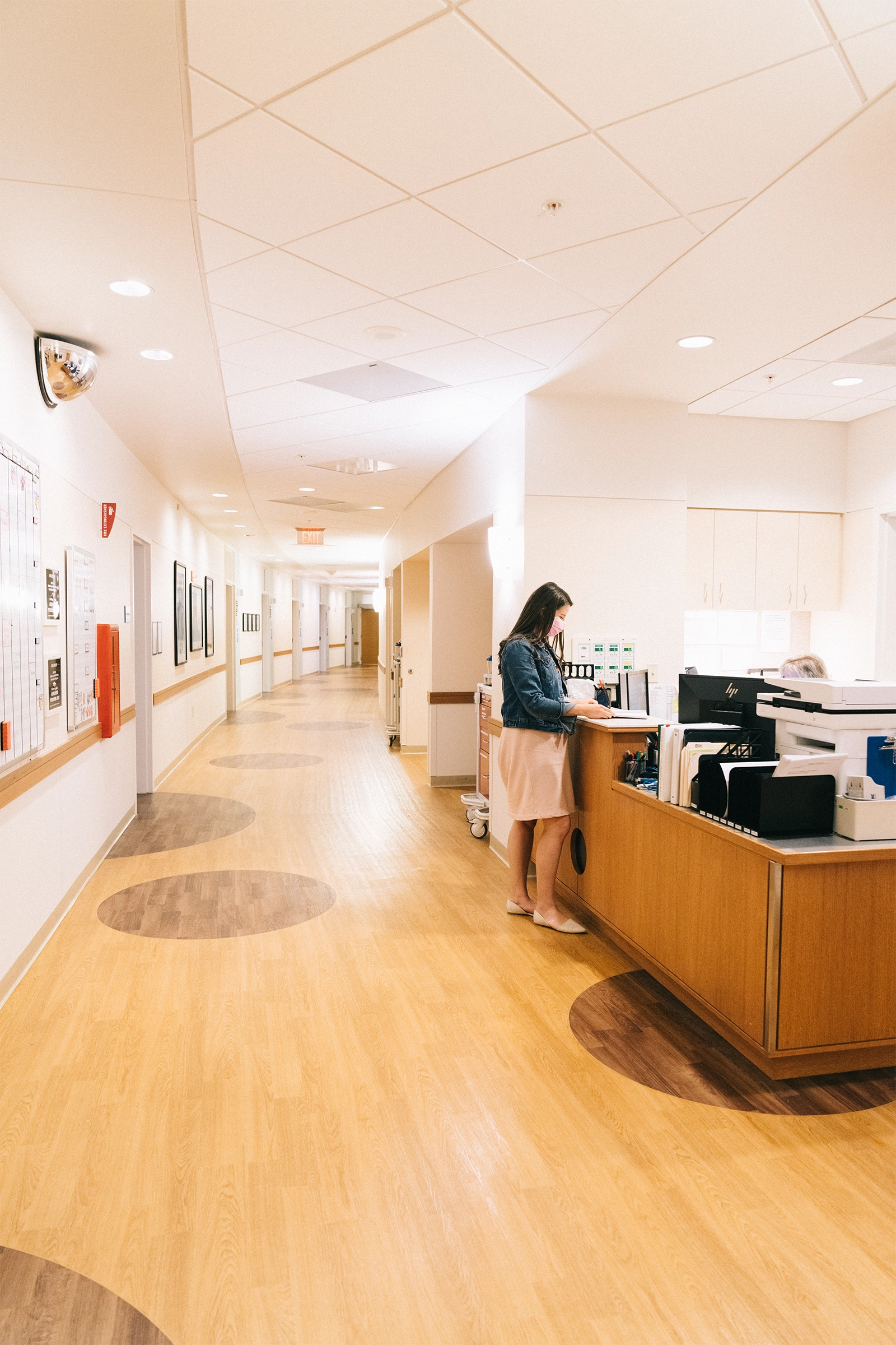 Woman standing at a hospital nurses' station in a brightly lit hallway with wood-patterned floors.