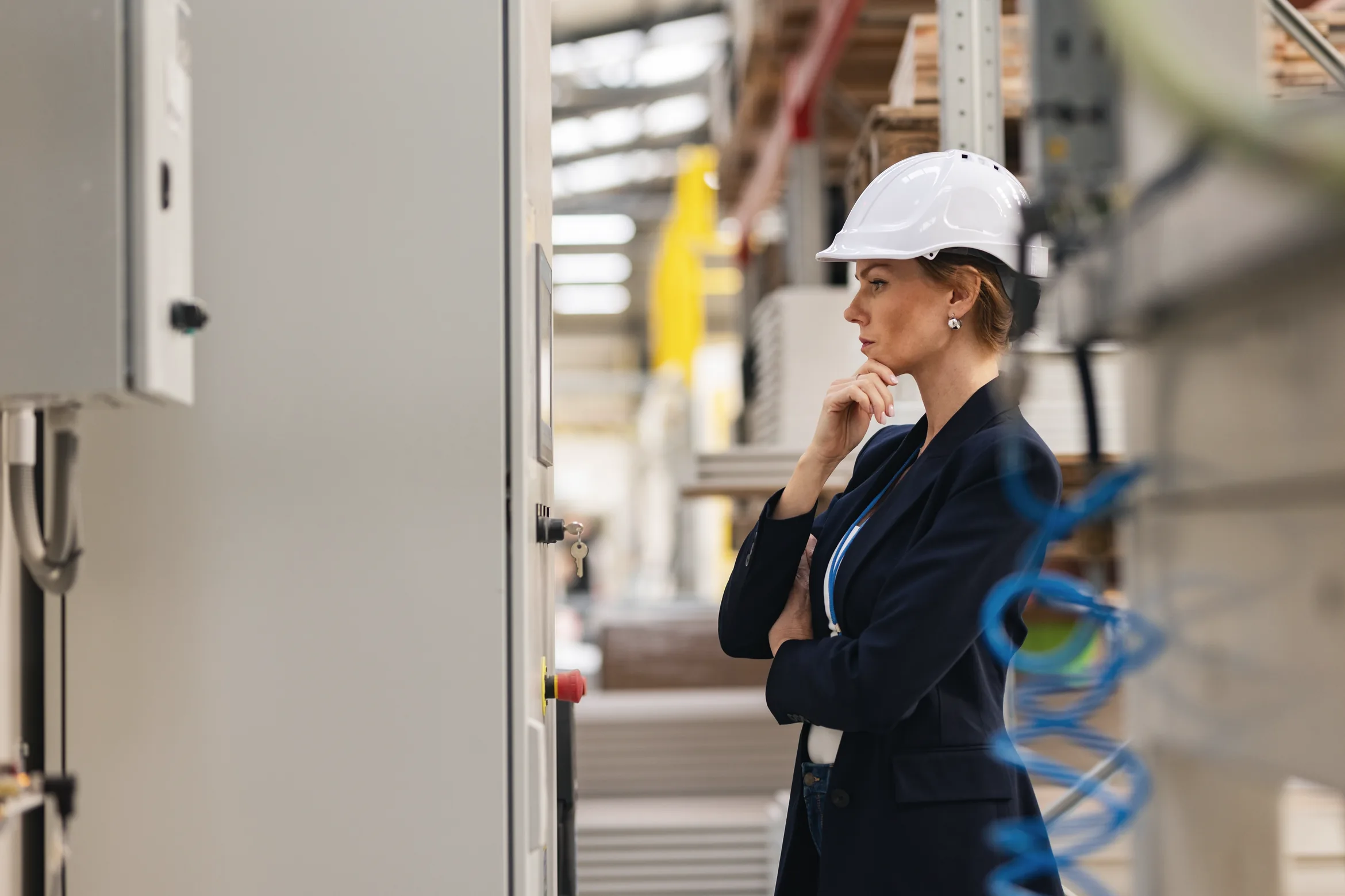 A manager in a hard hat and blazer, thinking, on the floor of a working factory