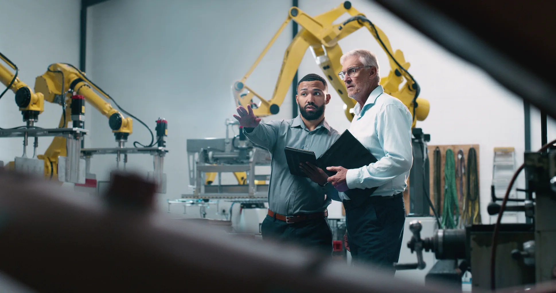 Two people talking over a tablet in front of robot arms on a factory floor